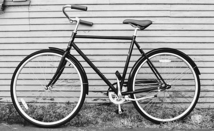 A black and white photo of an old-fashioned bicycle leaning against the outer wall of a house.