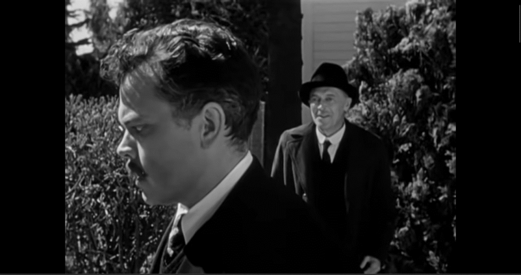A two-shot with one man in the foreground, looking off to the left. Another man is in the background to his right, looking at him.