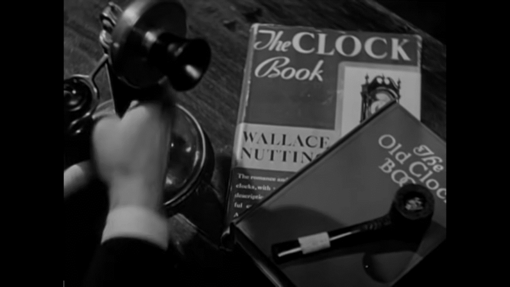 An extreme close-up of a man's hand reaching for a telephone. A book titled "The Clock Book" is on the desk next to the telephone.